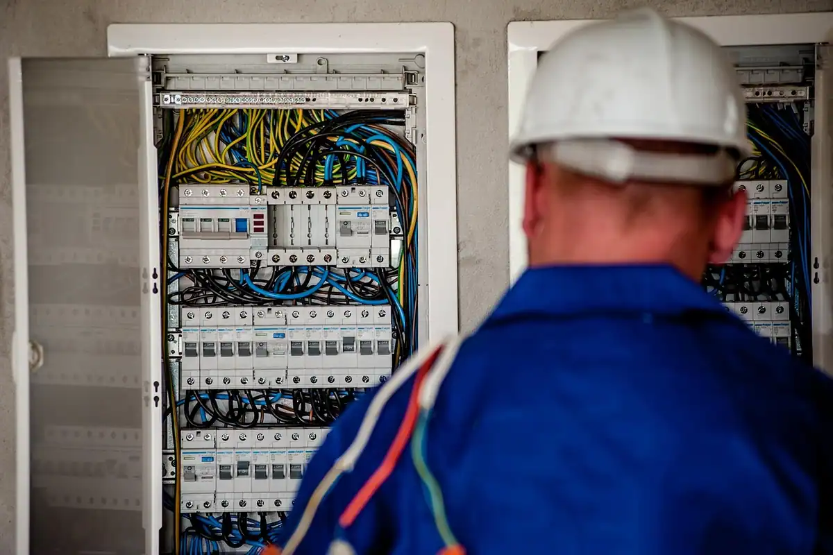 Electrician în cască albă lângă un tablou electric cu multe cabluri